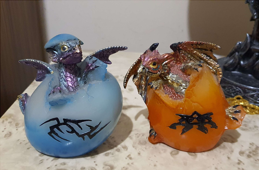 Two small statues of baby dragons hatching from the eggs. The left one is blue, and right is orange.